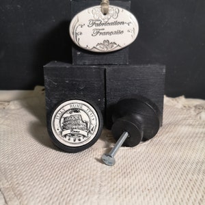 May include: Black cabinet knob with a white ceramic insert featuring a black and white image of the Colosseum in Rome, Italy. The text "Italy Rome Italy" is printed around the image. The text "Rome" is printed below the image. The knob is on a black surface.