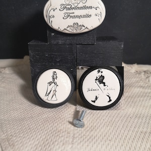 May include: Three ceramic drawer knobs with black bases. One knob features a woman in a dress, another features a man in a hat and walking stick, and the third knob has the text "Johnnie Walker" on it. The third knob also has a small black and white image of a man in a hat and walking stick. All three knobs have a white background with black details.