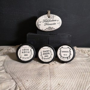 May include: A collection of decorative wine bottle stoppers. The stoppers have a black and white design with French text. The top stopper is oval-shaped and reads "Fabrication Française". The three round stoppers have wine names and years.