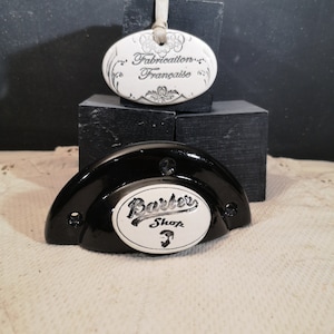 Peut inclure: Une enseigne de barbier en céramique noire avec un ovale blanc qui dit "Barber Shop" avec une image stylisée d'une barbe à l'intérieur de l'ovale. L'enseigne est sur un socle en bois noir. Une étiquette en céramique ovale blanche avec le texte "Fabrication Française" est sur un socle en bois noir.