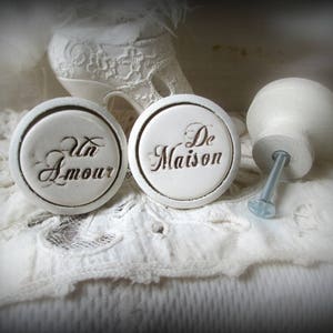 Peut inclure: Deux boutons en céramique blanche avec les mots "Un Amour" et "De Maison" écrits dessus. Les boutons sont sur un fond de dentelle blanche.