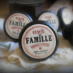 Könnte beinhalten: Eine Ansammlung schwarzer Schrankknöpfe mit weißen und roten Etiketten. Die Etiketten tragen den Text "esprit DE FAMILLE" und "MOMENTS A PARTAGER". Die Knöpfe sind auf einer weißen, strukturierten Oberfläche angeordnet.