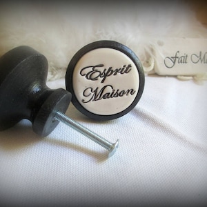 以下が含まれることがあります： 白地に「Esprit Maison」の文字が書かれた黒と白のセラミックノブ。ノブは黒い金属製のネジで取り付けられています。