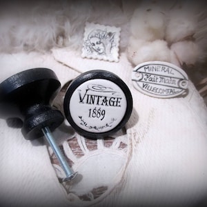 May include: Two black ceramic cabinet knobs with a vintage design. The knob on the left is plain, while the knob on the right has the words "Vintage 1889" written on it.