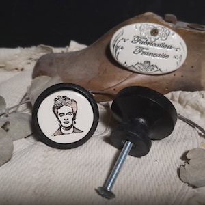 May include: Two ceramic drawer knobs, one with a black base and a white face featuring a black and white image of Frida Kahlo, and the other with a black base and a black face. The knobs are on a white fabric surface with a wooden shoe last in the background. The shoe last has a white oval label with the text "Fabrication Française".