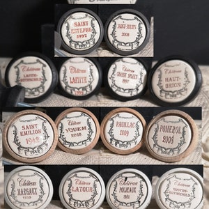 May include: A collection of vintage wine corks with various chateau names and years printed on them. The corks are round with a white background and black and red text. The image also includes a small ceramic tag that says "Fabrication Française".
