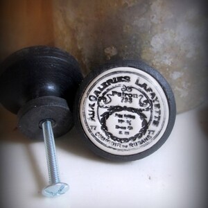May include: Two black ceramic drawer knobs with a white porcelain insert. The insert has a black and white design with the text "Aux Galeries Lafayette" and "Comptoir Rusans".