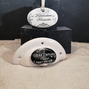 Peut inclure: Une poignée de porte en céramique blanche avec une étiquette ovale noire qui indique "Le prochain mot dans le cacao est CACAO PAYRAUD en Cuilles. Produit sans rival." La poignée a un trou sur le dessus et un petit trou sur le côté. Au-dessus de la poignée, une étiquette ovale en céramique blanche avec du texte noir qui indique "Fabrication Française".