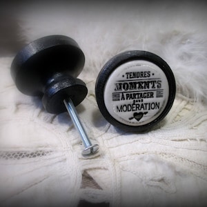 May include: Two black ceramic cabinet knobs with a silver screw. The knob on the right has a white ceramic insert with the text "Tendres Moments a partager sans moderation" and a small red heart.
