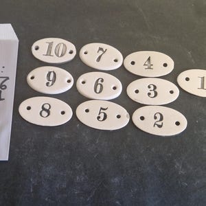 May include: A set of white ceramic oval-shaped tags with black numbers from 1 to 10. The tags are arranged in two rows. The numbers are written in a handwritten style. The tags are on a gray surface. The text on the paper bag reads "LOT 20: 10.9.8.7.6 5.4.3.2.1"