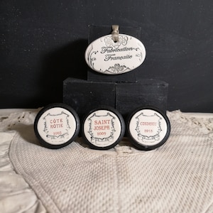 May include: Three round, black wine bottle stoppers with white labels. The labels have the names "Cote Rotie 2005", "Saint Joseph 2009", and "Condrieu 2015". A white oval ceramic tag with black lettering reads "Fabrication Française".