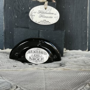 May include: Black ceramic bottle stopper with a white label that reads "Maison de Luxe". The stopper is sitting on a white lace doily. A small white oval tag with the text "Fabrication Française" is in the background.