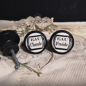 以下が含まれることがあります： 「EAU Chaude」と「EAU Froide」と書かれた黒と白のセラミックノブ2つ。ノブは白い布の背景の上に置かれています。