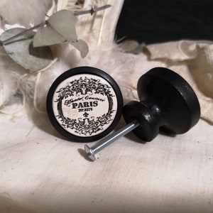 以下が含まれることがあります： 黒と白のセラミックノブに、花柄のデザインと「Hand Crafted Couture Paris NY 2273」の文字が書かれています。ノブは黒い木製のベースに銀色のネジで取り付けられています。