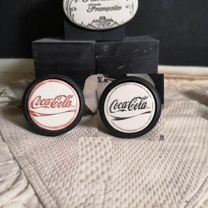 Può includere: Due tappi per bottiglia Coca-Cola in ceramica, uno con logo rosso e bianco e l'altro con logo nero e bianco. Entrambi i tappi hanno un bordo nero e sono esposti su una superficie nera.