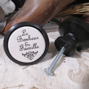 May include: Two black cabinet knobs. One knob has a white center with the text "Le Bonheur En Famille" in black script and a decorative flourish. The other knob is solid black with a screw attached.