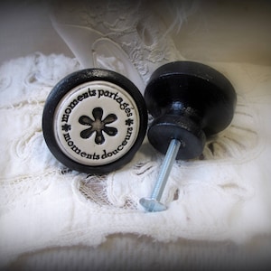 May include: Two black ceramic cabinet knobs with a white ceramic insert. The insert features a flower design and the text "moments partagés * moments douces".