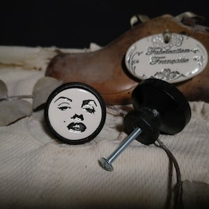 May include: Two ceramic cabinet knobs. One knob features a black and white image of Marilyn Monroe's face. The other knob is black.