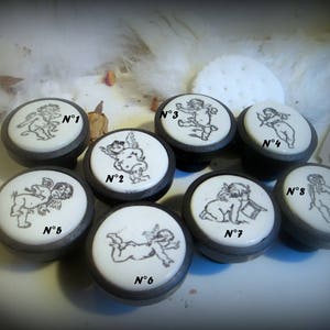 May include: Eight round ceramic knobs with black bases, each featuring a different black and white illustration of a cherub. The knobs are numbered 1 through 8.