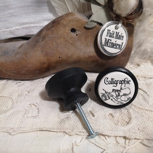 May include: Two black ceramic cabinet knobs. One knob has a white ceramic insert with the text "Calligraphie" and a black and white illustration. The other knob is plain black.