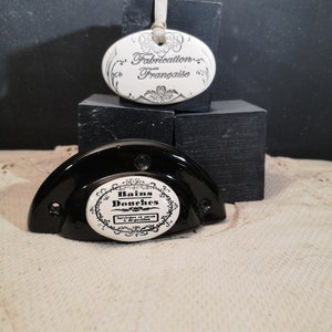 May include: A white ceramic oval tag with black text that reads "Fabrication Française" hanging from a white string. A black ceramic half-circle with a white ceramic oval tag that reads "Bains Douches" with black text.