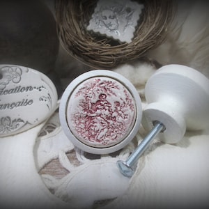 May include: Two ceramic cabinet knobs. One knob is white with a round base and a screw. The other knob is white with a red floral design.