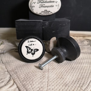 Puede incluir: Dos pomos de armario de cerámica negra con un diseño de mariposa. El pomo superior presenta el texto "Le battement d'aile..." en francés. El pomo inferior tiene un tornillo adjunto. Los pomos están sobre una superficie negra.