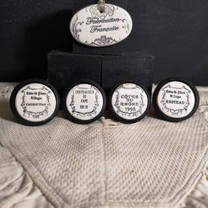 Peut inclure: Quatre bouchons de bouteille en céramique blanche avec inscription noire. Les bouchons représentent différentes régions viticoles françaises : Côtes du Rhône, Châteauneuf du Pape, Côtes du Roussillon Villages et Vacqueyras. Les bouchons sont disposés sur une surface noire.