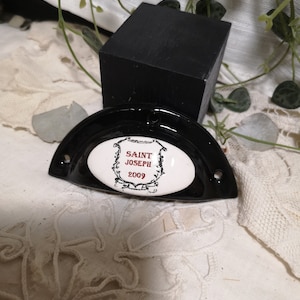 Peut inclure: Une poignée de tiroir noire, semi-circulaire, avec un centre ovale blanc. Le centre porte l'inscription "SAINT JOSEPH 2009" en rouge et noir. La poignée est posée sur un fond blanc texturé.