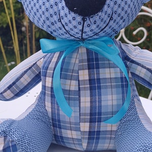 May include: A blue and white plaid teddy bear with a turquoise bow tie. The bear is sitting on a white surface with small holes.