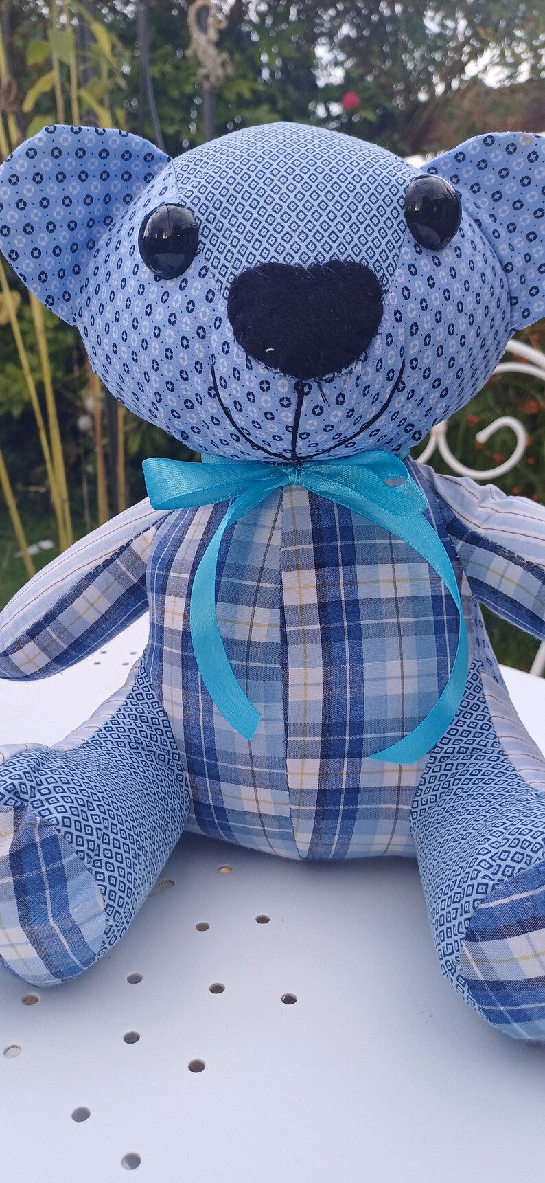 May include: A blue and white plaid teddy bear with a turquoise bow tie. The bear is sitting on a white surface with small holes.