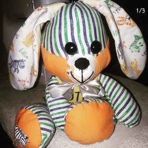 May include: A handmade stuffed bunny toy with green and blue striped fabric, orange fabric accents, and black button eyes. The bunny has a silver ribbon around its neck and is sitting on a white surface.