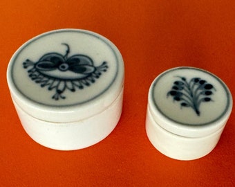 May include: Two white ceramic round lidded boxes with blue floral designs. The larger box has a more intricate design with a stylized flower, while the smaller box has a simple branch design.