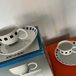 Może przedstawiać: Zestaw dwóch białych filiżanek do herbaty i spodków z czarnymi geometrycznymi wzorami. Filiżanki i spodki są zapakowane w niebieskie pudełko z nadrukiem "tête-à-tête" na przodzie.