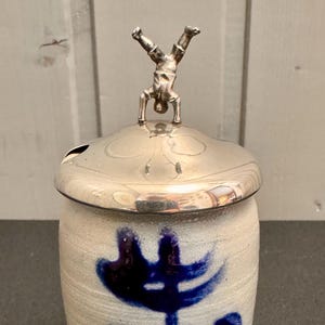 May include: A ceramic jar with a silver lid and a small figure on top. The jar is off-white with blue brushstrokes and the letters "ABB". The lid has a small opening for a spoon.