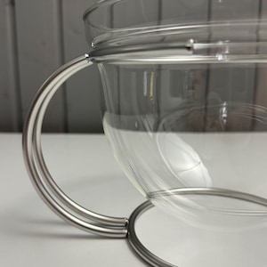 Puede incluir: Una taza de té de vidrio transparente con un asa y una base de acero inoxidable. El asa tiene un diseño curvo y semicircular. La taza está diseñada para contener bebidas calientes.