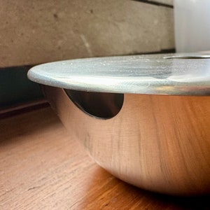 Peut inclure: Un cendrier en métal argenté avec un couvercle rond et un petit trou sur le côté. Le cendrier est posé sur une surface en bois.