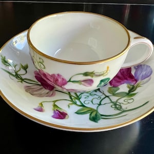 Pode incluir: Um conjunto de chávena e pires brancos com borda dourada. A chávena e o pires são decorados com desenhos florais roxos e rosa e vinhas verdes. A chávena tem uma pega curva.