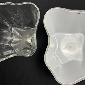 May include: Two decorative glass and white bowls with wavy edges. The clear glass bowl is on the left, and the white bowl is on the right. The bowls are empty and sit on a black surface.