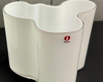 May include: A white glass vase with an organic, undulating, wave-like silhouette. The vessel features a smooth, opaque finish and a distinct red and white circular logo with the text iittala printed on the front surface of the glass structure.