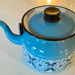 Puede incluir: Una tetera de esmalte azul con un diseño floral blanco y una tapa negra con un pomo de latón.