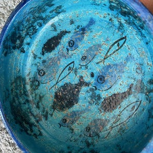May include: A round, turquoise ceramic bowl with a hand-painted fish design. The bowl's interior features a textured, mottled appearance with black and blue fish illustrations. The rim is a darker shade of blue.