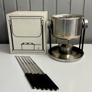 Può includere: Un set per fonduta vintage con una pentola in acciaio inossidabile, un supporto in metallo nero e forchette per fonduta. Le forchette hanno manici neri con punti colorati. Il set include una scatola con un disegno a tratto della pentola per fonduta.