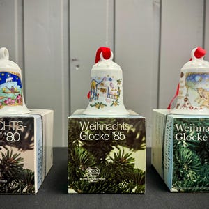 Op de afbeelding: Drie decoratieve kerstbellen op dozen. De bellen zijn wit met kleurrijke illustraties en rode linten. De dozen hebben de tekst "Weihnachts-Glocke" met de jaren 1980, 1985 en 1991.