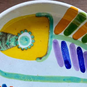 Peut inclure: Une assiette en céramique blanche avec un motif de poisson coloré. Le poisson est peint dans des tons de jaune, vert, bleu et violet. L'assiette est parfaite pour servir des en-cas ou des amuse-gueules.