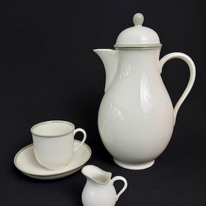 Könnte beinhalten: Ein weißes Keramik-Kaffeeservice mit grünem Rand. Das Set beinhaltet eine Kaffeekanne mit Deckel, einen Sahnekännchen, eine Tasse und eine Untertasse. Die Kaffeekanne ist etwa 25 cm hoch.
