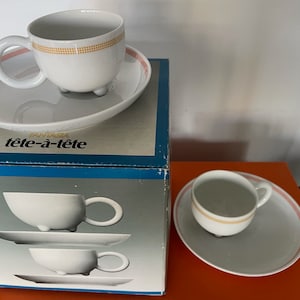 Może przedstawiać: Zestaw białych filiżanek do herbaty i spodków z złotą obwódką i różowymi akcentami. Filiżanki mają unikalny design z trzema małymi nóżkami. Pudełko zawiera napis "Fantasia tête-à-tête".