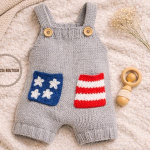 May include: A gray knitted baby romper with wooden button closures on the straps. The romper features two pockets, one with white stars on a blue background and the other with red and white stripes. A wooden rattle is next to the romper.