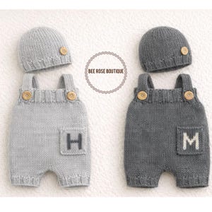 Puede incluir: Dos conjuntos de ropa de bebé de punto, cada uno con gorro y peto a juego. Uno es gris claro con la letra "H" en el bolsillo, y el otro es gris oscuro con la letra "M". Ambos conjuntos tienen detalles de botones de madera. El texto "BEE ROSE BOUTIQUE" está en un círculo.