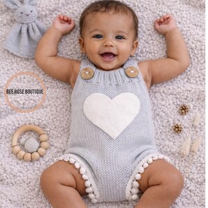 May include: A baby wearing a light blue knitted romper with a white heart applique and wooden button details. The romper has white pom-pom trim. A wooden teether and a stuffed bunny are nearby. The text "BEE ROSE BOUTIQUE" is in a circle.